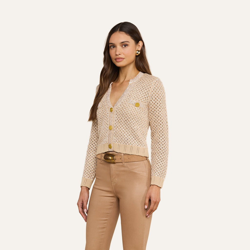 A woman wears the Blanca Sequin Cardigan, a neutral tan knit button-up with gold buttons, paired with light brown pants and a wide brown belt, standing against a plain beige background.