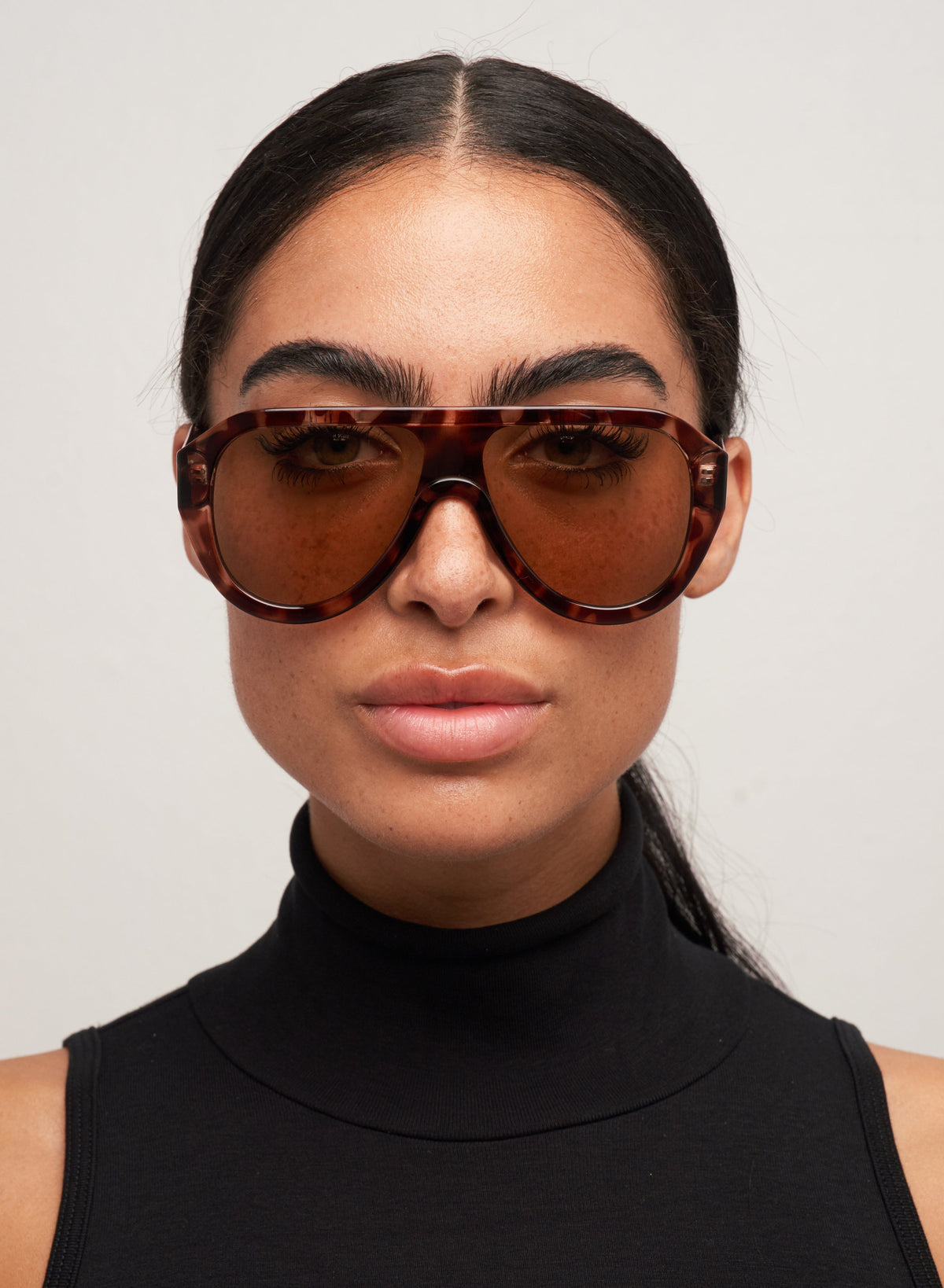 Otra Eyewear - Cameron Tort/Brown - Model wearing the Cameron sunglasses featuring a classic aviator silhouette crafted in tortoiseshell lightweight acetate with warm brown lenses in a front portrait shot against a light grey background. Effortless, refined, and designed for all-day wear.