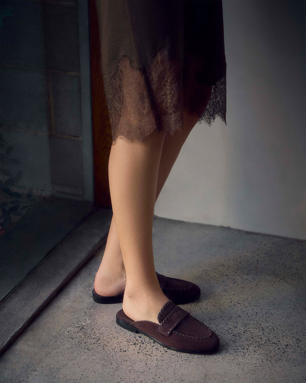 A person wearing dark brown slip-on loafers with a fringed strap detail, standing on a textured concrete floor next to a doorway.